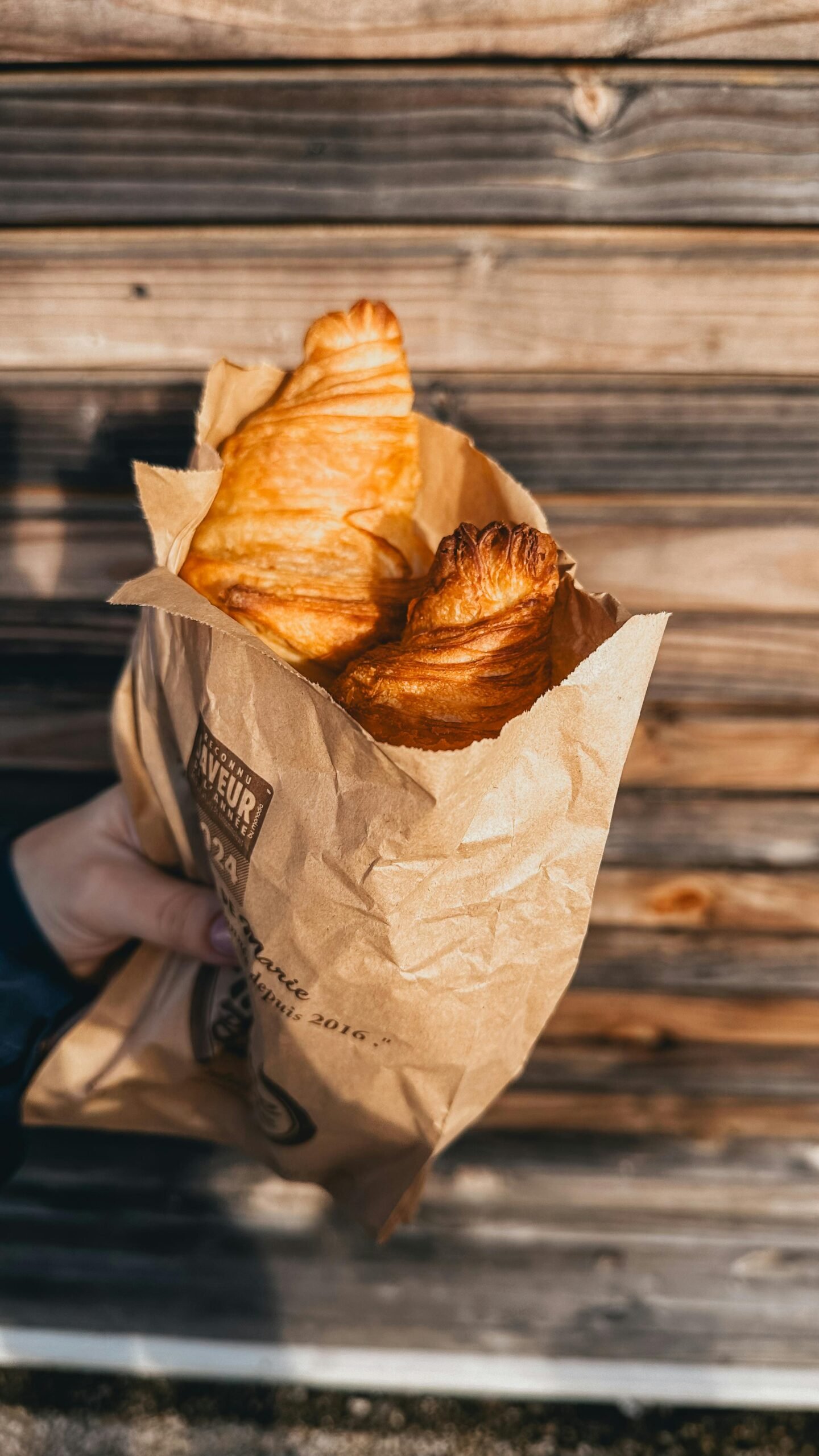 GÎTE ET CHAMBRE A person holding fresh croissants in a paper bag against a wooden background.